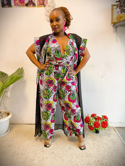 Woman wearing a colorful floral jumpsuit standing indoors with a plant and flowers in the background.
