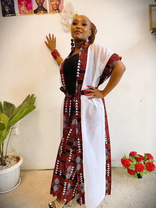 Woman in a patterned dress with a white overlay standing indoors with decor elements.